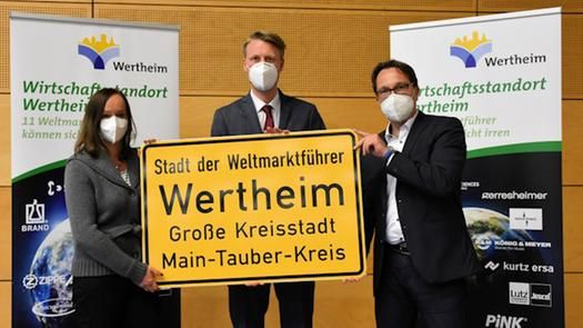 Ulrike Müller, Oberbürgermeister Herrera Torrez und Jürgen Strahlheim halten ein Ortsschild "Stadt der Weltmarktführer" in die Kamera