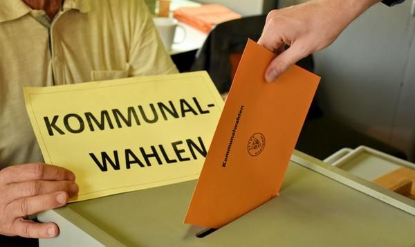 In eine Wahlurne mit der Aufschrift "Kommunalwahl" wird ein Wahlschein in orangefarbenen Umschlag gesteckt. 