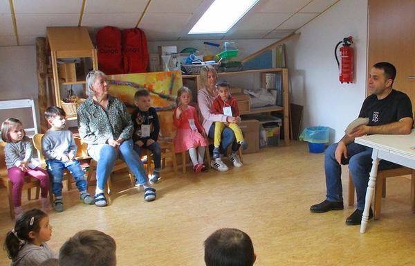 Kindergartenkinder, ihre Erziherinnen und ein Papa sitzen anlässlich des "Tag der Muttersprache" in einem Stuhlkreis, während der Papa den Kindern etwas in seiner Muttersprache beibrigt. 