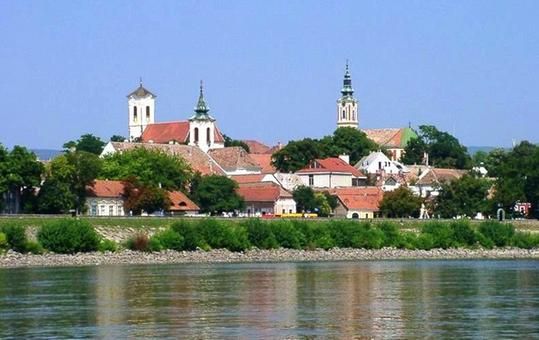 Aufnahme der Stadt Szentendre in Ungarn. Im Vordergrund ist ein Fluss zu sehen, in der Ferne sieht man drei Kirchtürme und einige Häuser