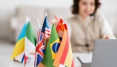 Symbolbild Ausländerbehörde: Viele kleine Flaggen verschiedener Länder stehen bei einer Frau auf dem Schreibtisch. Diese ist im Hintergrund mit Laptop und Headset zu sehen.