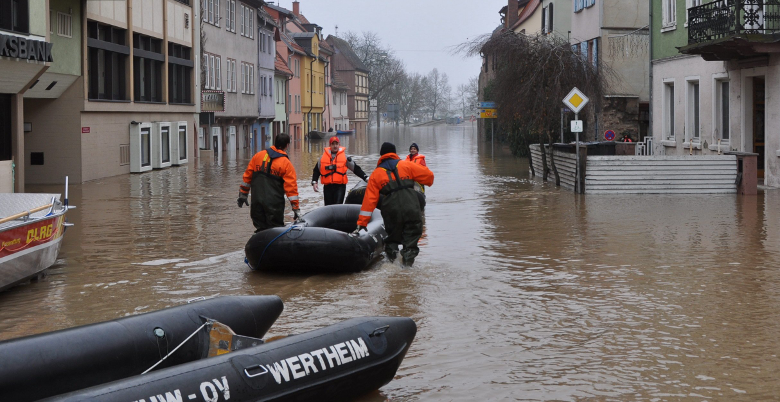 Die überflutete Wertheimer Innenstadt. Ebenfalls zu sehen sind Hochwasserboote und vier Männer in orangenen Jacken, die eines der Hochwasserboote durch die Fluten  schieben.