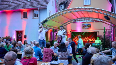 Viele Menschen sitzen bei stimmungsvoller Beleuchtung auf Bierbänken und hören einer Band zu. Das Foto stammt vom Wertheimer Altstadtfest.