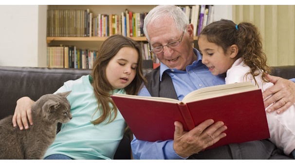 Ein älterer Herr liest zwei Mädchen und einer Katze etwas aus einem roten Buch vor.
