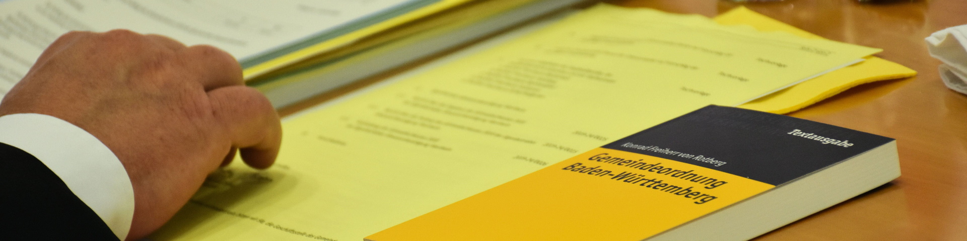 Symboldbild Kommunalpolitik: Auf einem Tisch liegen die gelben Vorlagen und die Gemeindeordnung. Es ist zudem eine Männerhand zu sehen.