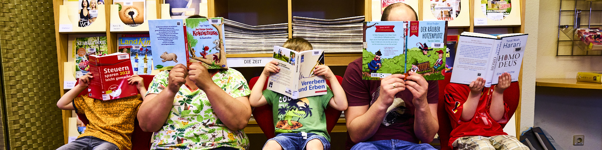 Drei Kinder und zwei Erwachsene sitzen in der Bücherei. Ihre Gesichter verdecken sie mit aufgeschlagenen Büchern. 