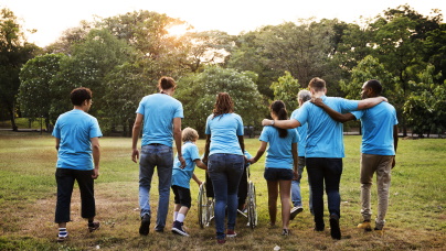 Symbolbild: Acht Personen, Männer, Frauen und Kinder in blauen T-Shirts, sind von hinten zu sehen. Sie befinden sich draußen auf einer Wiese. Eine Person sitzt im Rollstuhl und wird geschoben. 